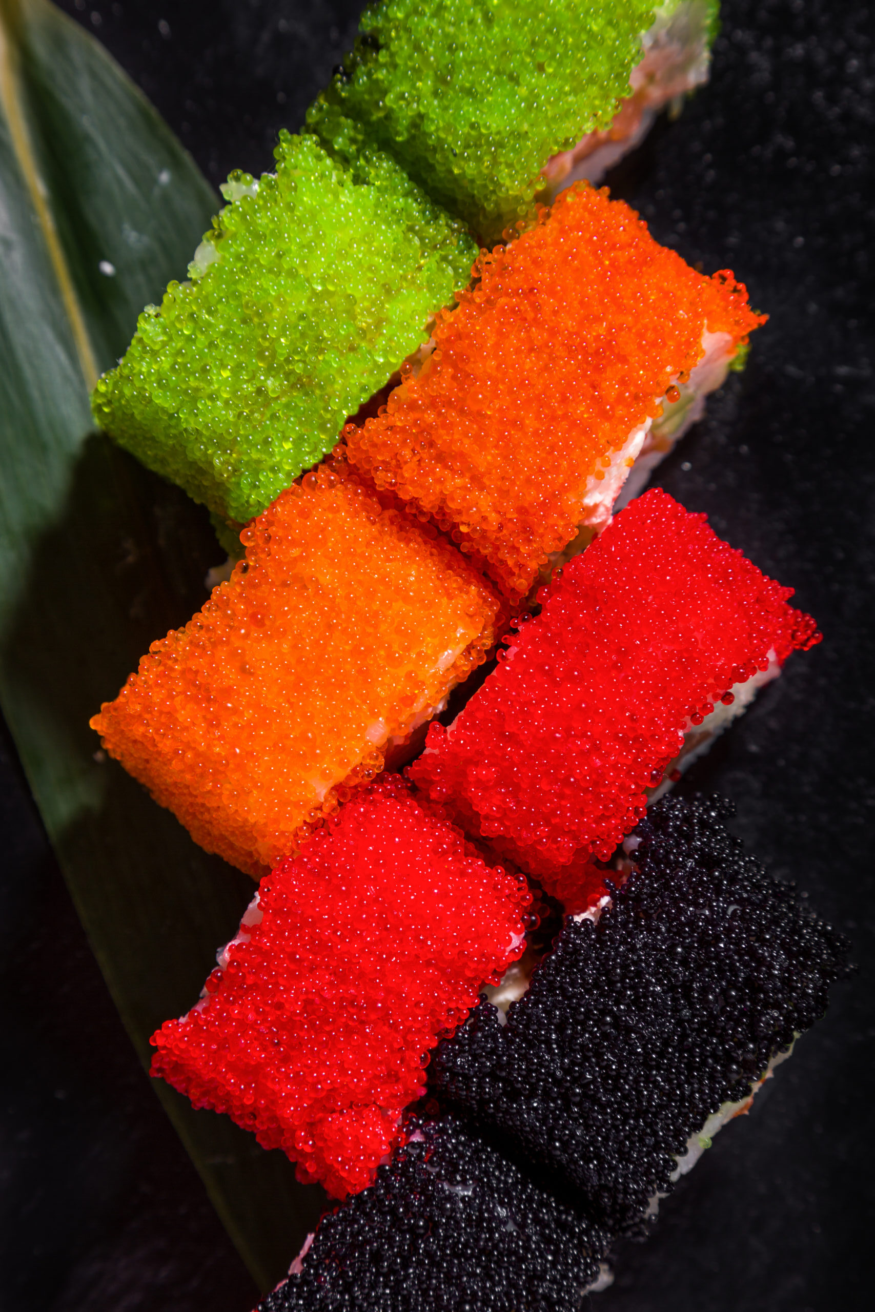 Rolls with rice, cream cheese, salmon, with red, black, orange and green tobiko caviar on a leaf on a black background