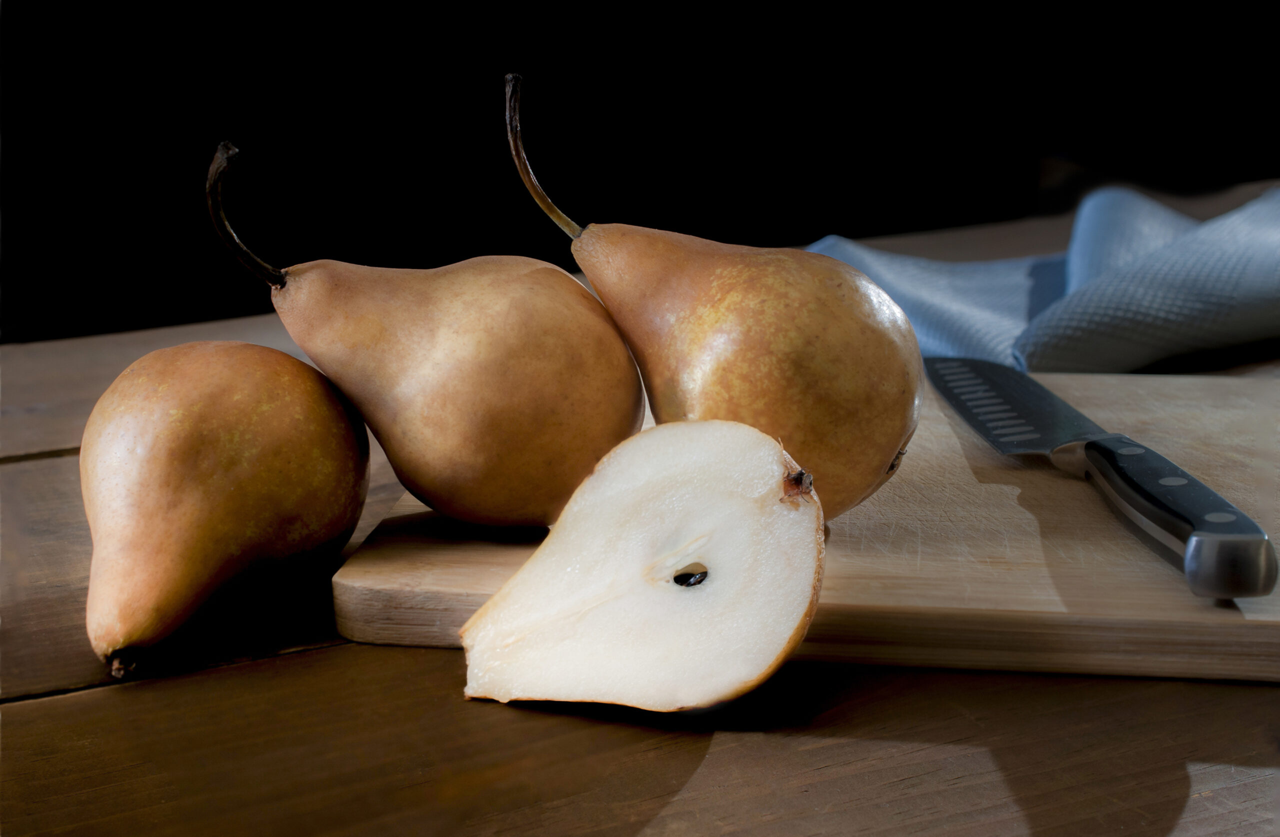 Bosc pears or brown pears on a cutting board with a knife on the side and a blue napkin at the back in a black background. Rim light or backlight